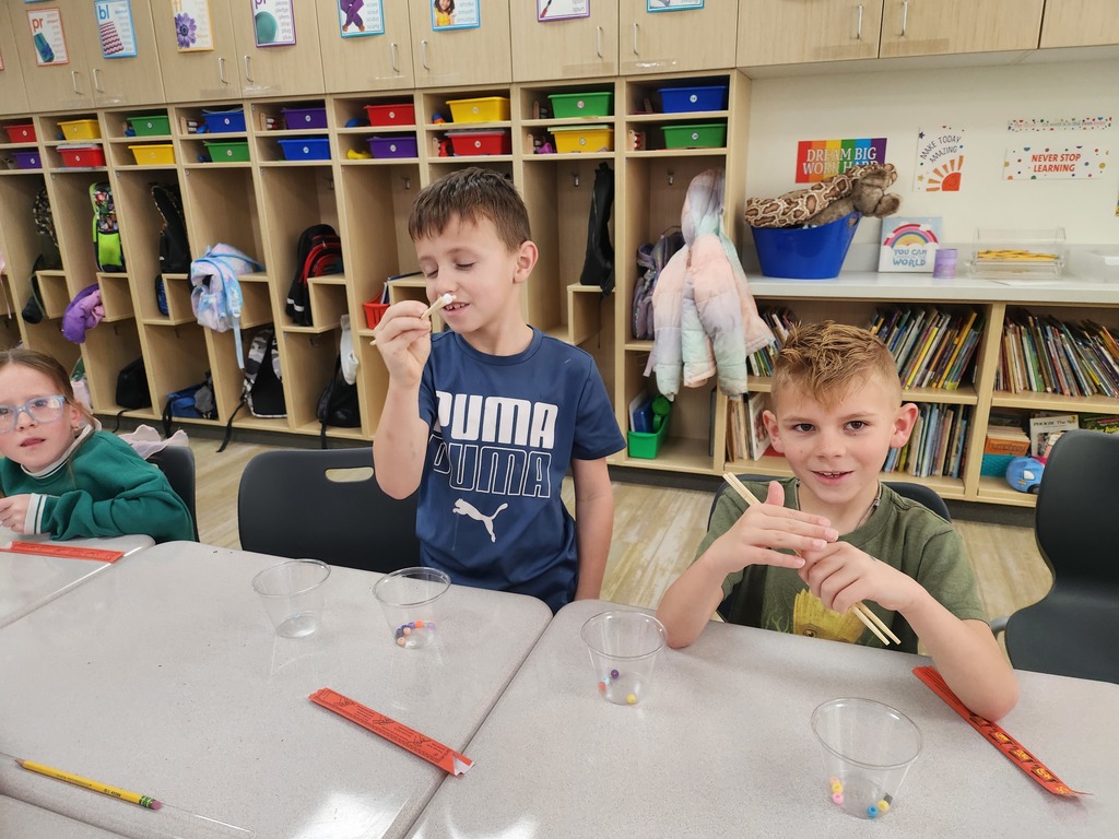 Three elementary students sitting at a classroom table practicing using wooden chopsticks to pick up colorful beads from clear plastic cups. One boy in a blue Puma shirt holds a bead up to inspect it. In the background, wooden cubbies hold colorful bins and backpacks. Visible text on the wall includes: "DREAM BIG WORK HARD," "MAKE TODAY AMAZING," "YOU CAN WORLD," and "NEVER STOP LEARNING."