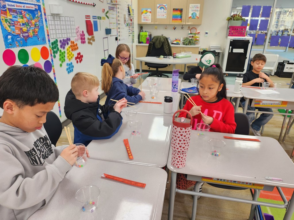 A group of students at their desks in a classroom focusing on a fine motor activity with chopsticks and beads. A girl in a bright red sweatshirt concentrates on her cup. On the wall, a map of the "United States of America" is visible alongside educational posters. A tall water bottle with a red heart pattern sits on the desk. Visible text on cubby labels and posters includes: "KIND THINGS," "SOMETHING," and "CLASS."