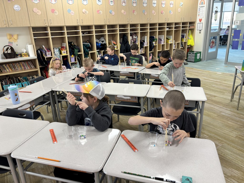 A classroom scene from a high angle showing students at their desks using chopsticks to pick up beads. The back wall is lined with light wood lockers/cubbies featuring rainbow-themed circular labels for the months of the year, such as "January," "February," and "March." One student wears a hat with a rainbow brim. A green crate is visible in the corner of the room.