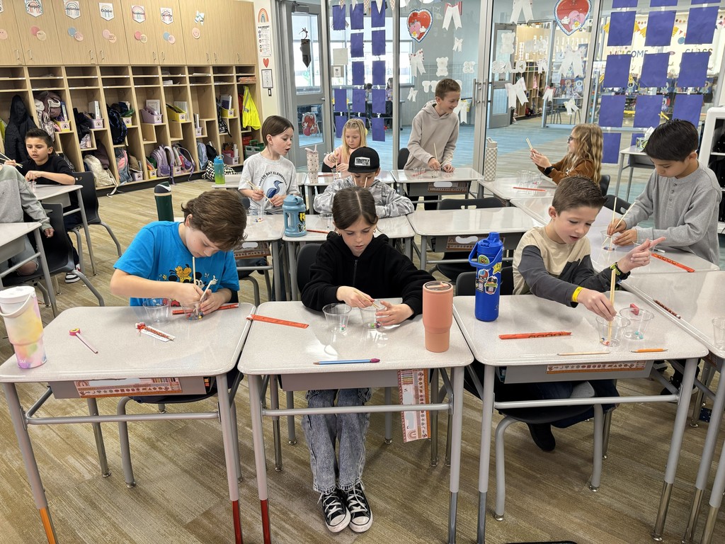 An overhead wide shot of a classroom where a dozen students are seated at desks arranged in rows, all participating in a chopstick activity. The room features light wood cabinetry and blue paper displays on a glass partition. One student wears a shirt that says "HAVEN BAY ELEMENTARY."