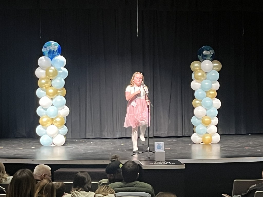 a girl in a pink dress standing on a stage with balloons and a microphone at the storytelling festival