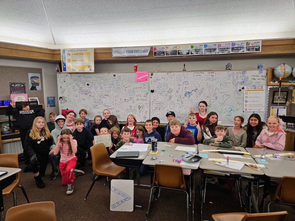 A wide shot of a diverse classroom of elementary students at Lomond View Elementary. They are gathered in front of a large whiteboard that is completely covered in hand-drawn hearts and messages of love and kindness. The students are smiling, making peace signs, and posing playfully. The classroom is filled with desks, educational posters, and Valentine's Day treats.