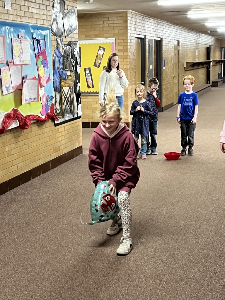 A series of photos showing first-grade students at Lomond View Elementary celebrating Valentine's Day. Activities include students sitting on a colorful rug making heart-shaped bead crafts, playing Valentine-themed Bingo at small tables, decorating and eating heart-shaped cookies with pink frosting, and participating in a "balloon waddle" relay race in the school hallway. Teachers and parent volunteers are seen guiding the various craft and game stations in a decorated classroom.