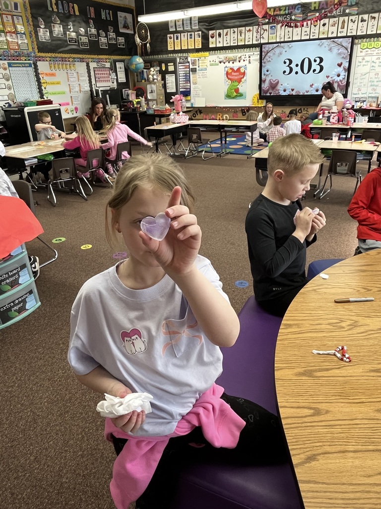 A series of photos showing first-grade students at Lomond View Elementary celebrating Valentine's Day. Activities include students sitting on a colorful rug making heart-shaped bead crafts, playing Valentine-themed Bingo at small tables, decorating and eating heart-shaped cookies with pink frosting, and participating in a "balloon waddle" relay race in the school hallway. Teachers and parent volunteers are seen guiding the various craft and game stations in a decorated classroom.