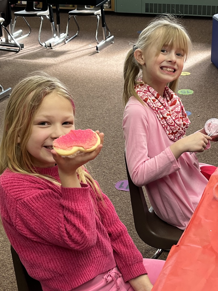 A series of photos showing first-grade students at Lomond View Elementary celebrating Valentine's Day. Activities include students sitting on a colorful rug making heart-shaped bead crafts, playing Valentine-themed Bingo at small tables, decorating and eating heart-shaped cookies with pink frosting, and participating in a "balloon waddle" relay race in the school hallway. Teachers and parent volunteers are seen guiding the various craft and game stations in a decorated classroom.