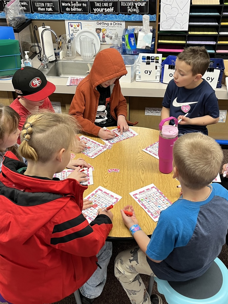 A series of photos showing first-grade students at Lomond View Elementary celebrating Valentine's Day. Activities include students sitting on a colorful rug making heart-shaped bead crafts, playing Valentine-themed Bingo at small tables, decorating and eating heart-shaped cookies with pink frosting, and participating in a "balloon waddle" relay race in the school hallway. Teachers and parent volunteers are seen guiding the various craft and game stations in a decorated classroom.