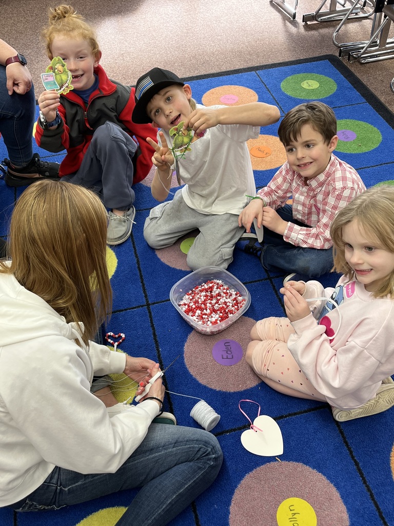 A series of photos showing first-grade students at Lomond View Elementary celebrating Valentine's Day. Activities include students sitting on a colorful rug making heart-shaped bead crafts, playing Valentine-themed Bingo at small tables, decorating and eating heart-shaped cookies with pink frosting, and participating in a "balloon waddle" relay race in the school hallway. Teachers and parent volunteers are seen guiding the various craft and game stations in a decorated classroom.