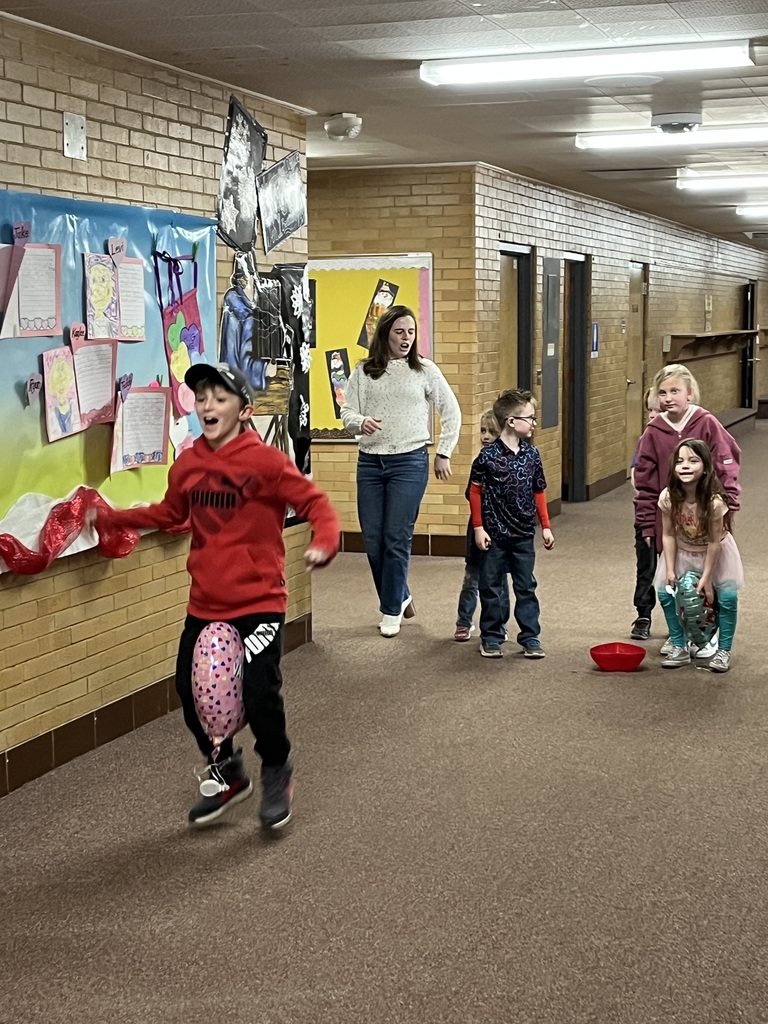 A series of photos showing first-grade students at Lomond View Elementary celebrating Valentine's Day. Activities include students sitting on a colorful rug making heart-shaped bead crafts, playing Valentine-themed Bingo at small tables, decorating and eating heart-shaped cookies with pink frosting, and participating in a "balloon waddle" relay race in the school hallway. Teachers and parent volunteers are seen guiding the various craft and game stations in a decorated classroom.
