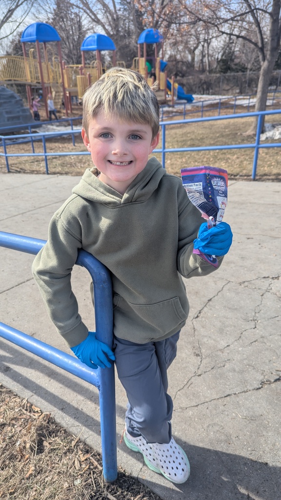 A young boy in an olive green hoodie and blue gloves leans against a blue playground railing. He is smiling and holding up an empty candy wrapper he collected, with the school playground equipment visible behind him.