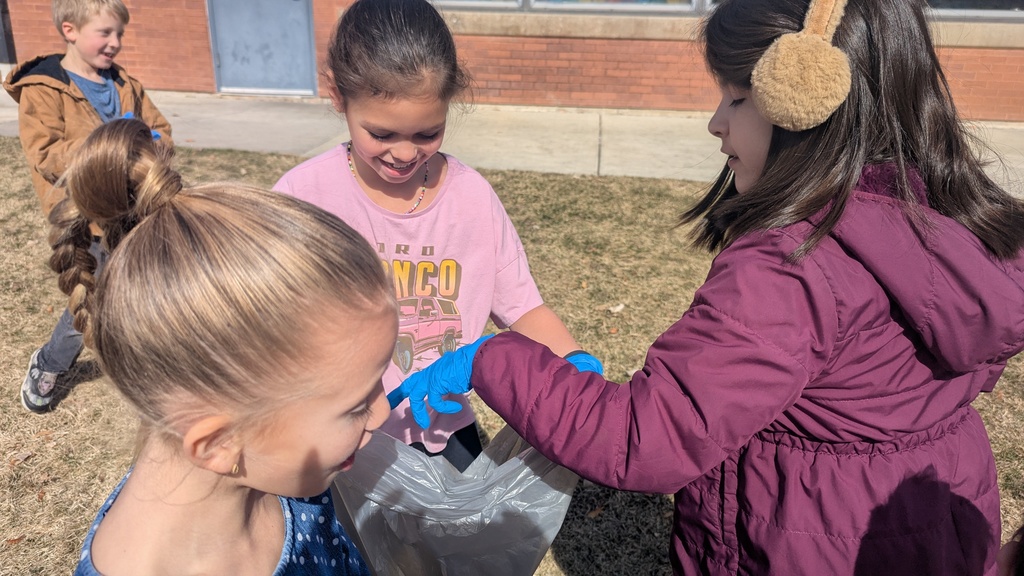 A close-up shot of three young girls smiling as they work together. One girl in a purple jacket and another in a pink "Bronco" shirt are carefully placing a piece of trash into a grey collection bag held by a classmate.