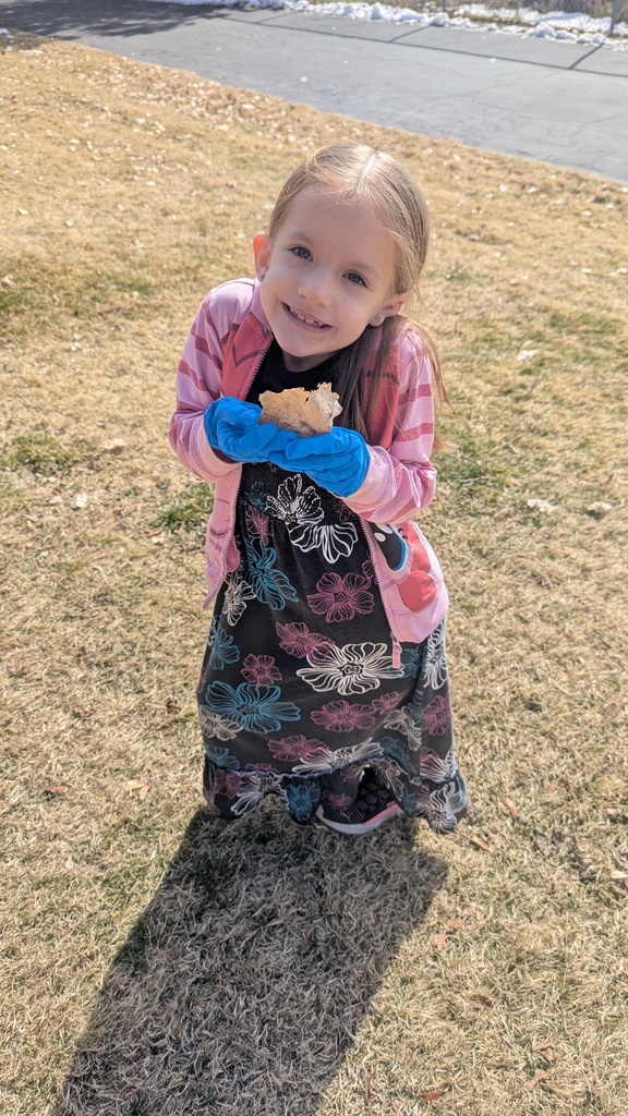 A young girl in a floral dress and pink zip-up hoodie smiles brightly at the camera. She is wearing blue protective gloves and proudly holding up a piece of litter she found during the cleanup.