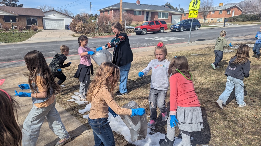 A large group of elementary-aged students and a female teacher wearing bright blue gloves, working together to pick up litter on a grassy area near a school crossing sign. They are filling large grey plastic bags on a sunny day.