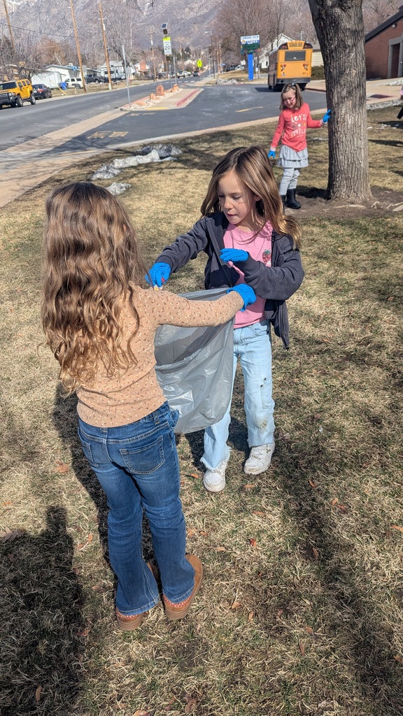Two girls on a lawn near the school bus zone; one girl in a tan sweater holds open a grey trash bag while her friend in a grey hoodie and light blue jeans places litter inside. A school bus is visible in the background.