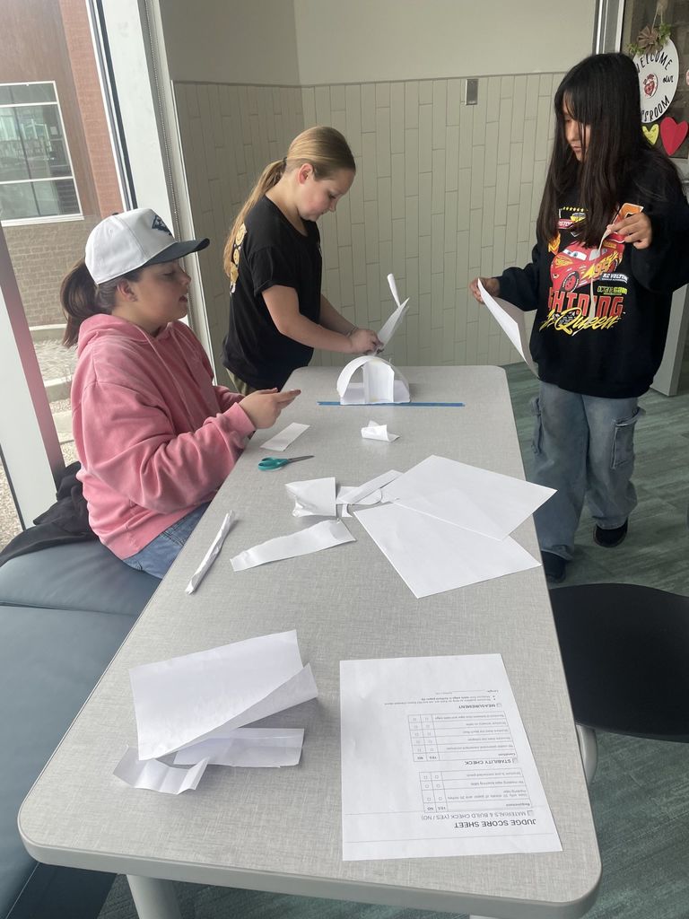 Three girls are focused on building a white paper arch structure on a long table near a window. One girl wears a pink hoodie and a white baseball cap, another is in a black t-shirt, and the third wears a black hoodie with a "Lightning McQueen" graphic and the words "Shining Queen." A "Judge Score Sheet" is prominently displayed on the table.