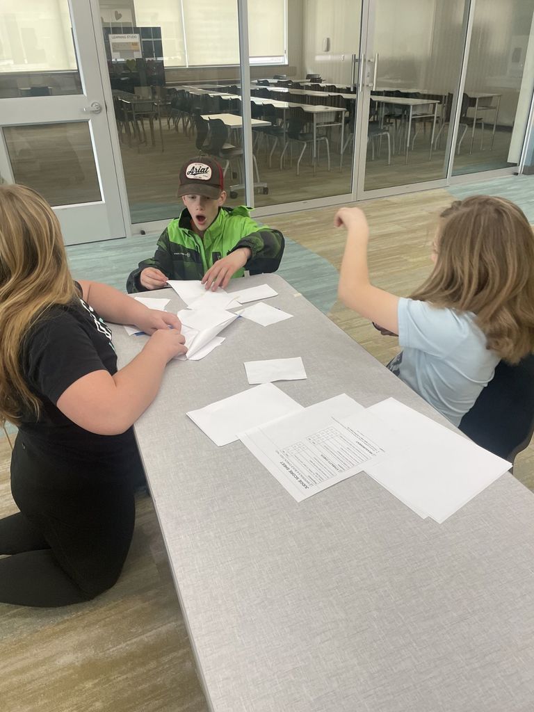 A boy in a bright green and black jacket with an "Ariat" hat sits at a table, looking surprised with his mouth open while working with paper. Two other students are partially visible on either side, helping fold and organize paper strips. A "Judge Score Sheet" lies on the table in front of them.