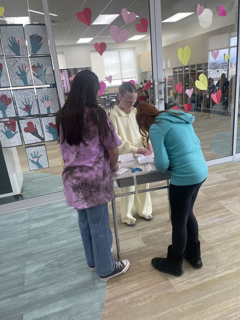 Three students are huddled over a small desk, collaborating on a paper craft. One student wears a purple tie-dye shirt, another a pale yellow tracksuit, and the third a bright turquoise hoodie. Blue scissors and scraps of white paper are on the desk. In the background, a glass wall is decorated with red, pink, and yellow paper hearts and "handprint" art.