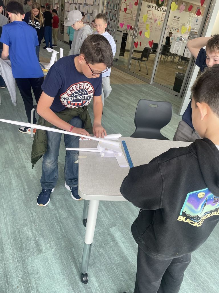 A boy in an "American Chevrolet" t-shirt and glasses leans over a table, carefully assembling a long, pointed paper structure. Other students are visible in the background of the open hallway. The structure is being built over a blue line of tape on the table surface. A boy in a black "Bucket Squad" hoodie stands in the foreground.