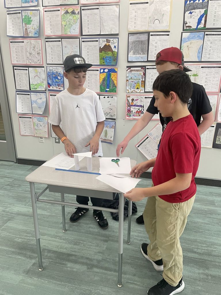Three boys work on a STEM project involving a small paper bridge structure. One boy in a white Jordan t-shirt and grey baseball cap looks down at the structure. Another boy in a red t-shirt holds a piece of paper. The background wall is covered in student drawings of ecosystems, including volcanoes and waterfalls, under the heading "Interactions with Earth's Systems."