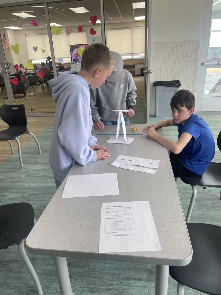 Three students work on a paper structure that resembles a small table or goalpost. A boy in a light blue hoodie leans in to observe the build. On the table, a "Judge Score Sheet" is visible with sections titled "MATERIALS & BUILD CHECK," "STABILITY CHECK," and "MEASUREMENT." In the background, a glass door is decorated with colorful paper hearts.
