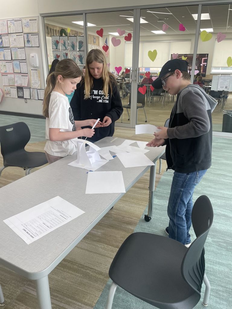 Three students, two girls and a boy in a black baseball cap, stand around a light gray table working on a paper engineering project. A white "Judge Score Sheet" with a table of criteria sits in the foreground. They are using strips of white paper and tape to build a structure. The girl in the middle wears a black hoodie that reads, "I'M COLD. I'm Always Cold... ME 7/24."