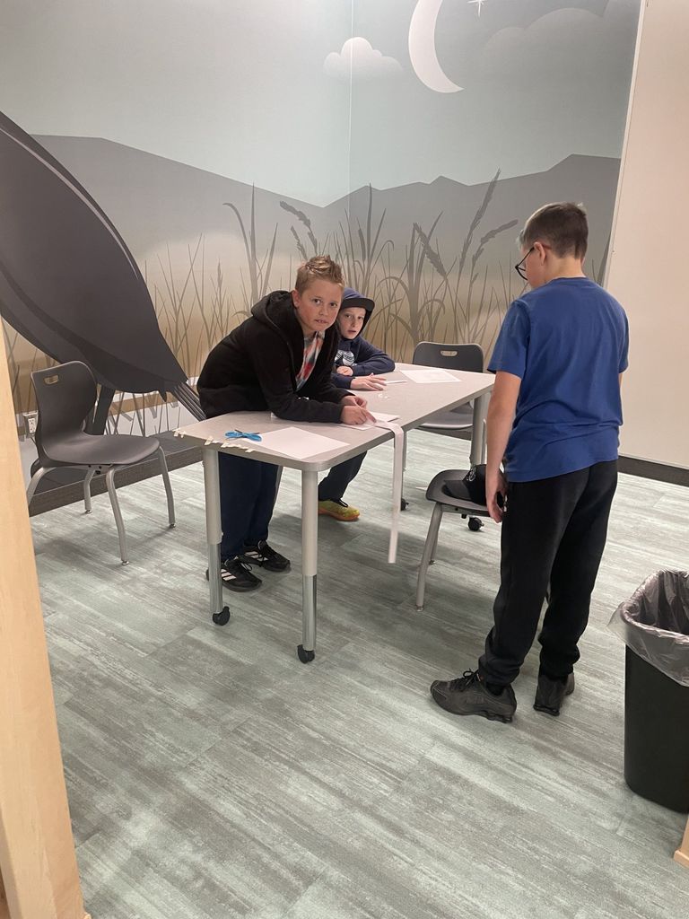 Three boys work at a table in a room featuring a large mural of a night sky with a crescent moon and tall grass. One boy in a black jacket leans on the table, looking toward the camera, while another in a blue t-shirt stands nearby. A long, thin strip of white paper hangs off the edge of the table next to a pair of blue scissors.