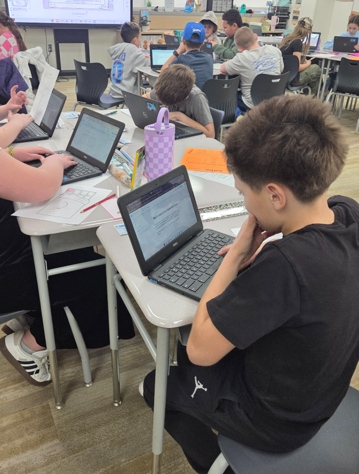 A student in a black t-shirt sits at a desk, typing on a Dell Chromebook laptop. The screen shows a Google Doc titled "What it means to be a Haven Bay Heron." Other students in the background are also working on laptops, and a purple checkered water bottle sits on the desk nearby. A large digital whiteboard is visible at the front of the room showing a lesson.