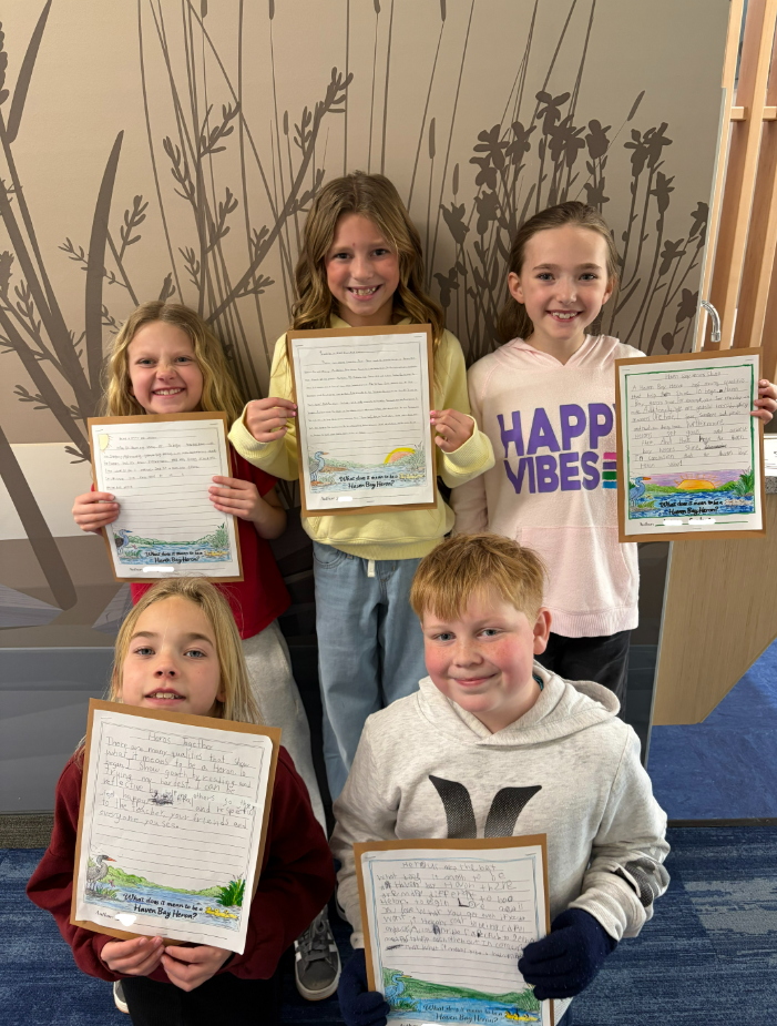 Five smiling elementary school students stand together against a wall with a nature-themed mural, proudly holding up their handwritten essays titled "What does it mean to be a Haven Bay Heron?" Each paper features a colorful illustration of a heron in a marsh at the bottom. The essays include phrases like "Herons Together," "Herons are the best," and "I show growth by reaching and trying my hardest."
