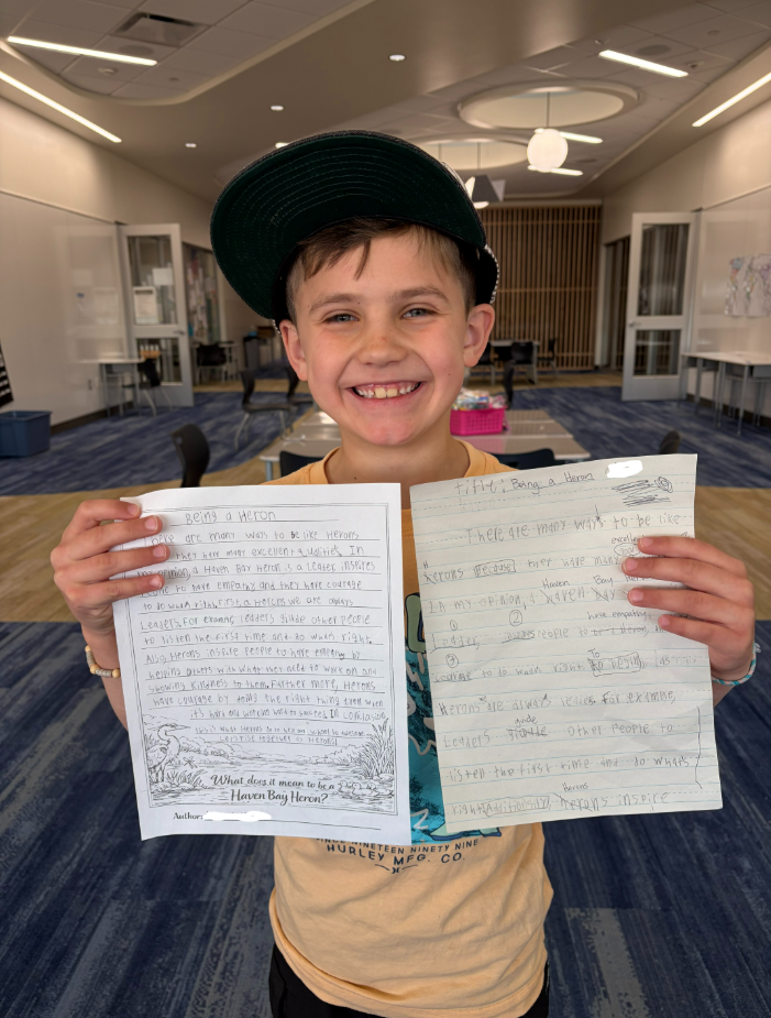 A young boy wearing a green baseball cap and a yellow "Hurley MFG. CO." t-shirt smiles broadly while holding two versions of his essay titled "Being a Heron." One is a rough draft with handwritten edits, and the other is a final printed copy. The text reads: "There are many ways to be like Herons... In my opinion, a Haven Bay Heron is a leader, inspires people to have empathy, and they have courage to do what's right." At the bottom, a prompt asks: "What does it mean to be a Haven Bay Heron?"