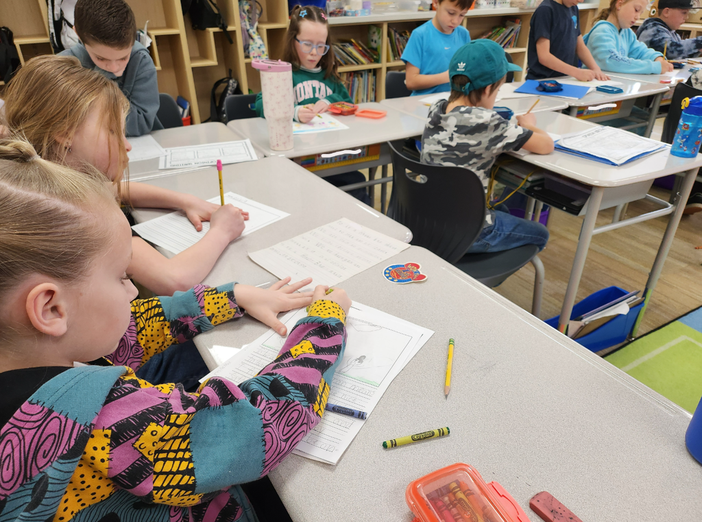 A classroom scene showing several children sitting at white desks, diligently working on their writing projects. In the foreground, a student in a colorful "The Nightmare Before Christmas" patterned hoodie writes on a worksheet. Another student’s paper is visible with the handwritten text: "To be a Heron you have to work and show kindness. We are herons..." There are crayons, erasers, and a "Montana" water bottle on the desks.