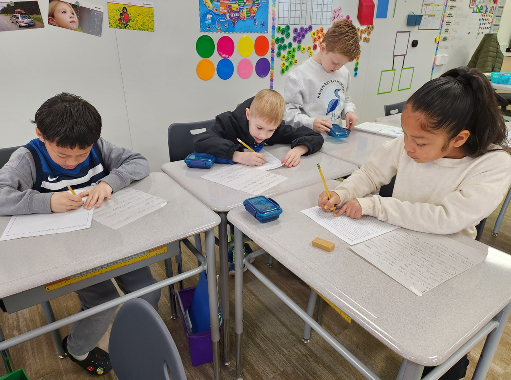 our elementary students sit at a cluster of gray desks in a classroom, focused on writing their essays with pencils. One student wears a grey sweatshirt with a blue heron logo that says "Haven Bay Elementary." The classroom wall in the background is decorated with a map of the United States, colorful circular dots, and a "Check In" chart.