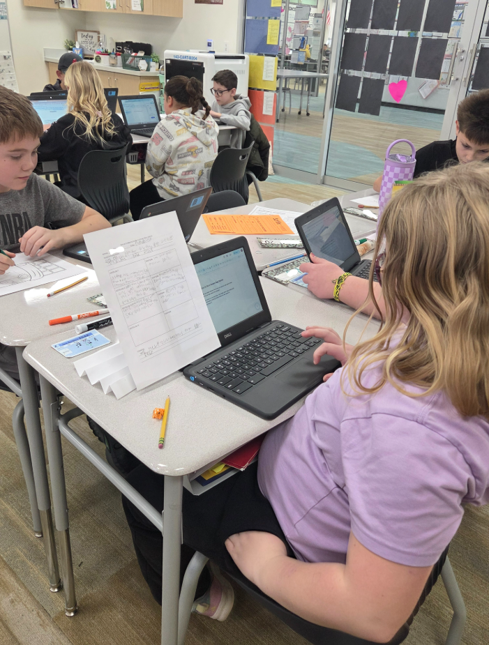 An over-the-shoulder view of a student with blonde hair working on a Dell laptop in a classroom. Next to the laptop is a handwritten graphic organizer titled "Being a Haven Bay Heron" with sections for "Opinion," "Reason 1," "Reason 2," and "Closing." The closing section says: "We help ourselves to be better." Other students and a "143-CART-B204" charging station are visible in the background.