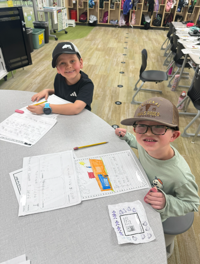 Two young boys sit at a curved gray table, smiling at the camera. One boy wears a black hat with a white logo, and the other wears a brown "National Finals Rodeo" hat and glasses. On the table are their worksheets; one is an "Opinion" organizer titled "Being a Haven Bay Heron" where the student wrote, "I am paying attention." The other student has a colorful drawing of a building with the text: "I am a Haven Bay Heron... I am a Heron."