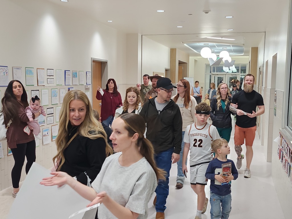 A crowded school hallway filled with parents and children walking during a literacy event. The walls are lined with student work and colorful displays. A boy in a white "Prospects 22" basketball jersey walks through the center of the frame.