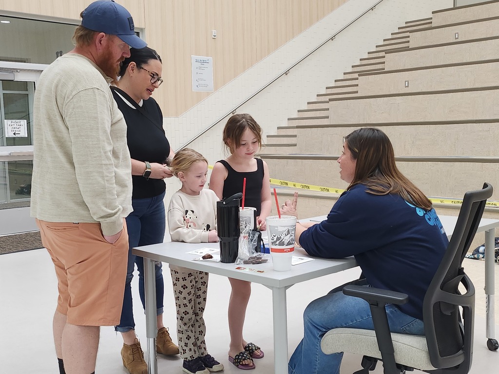 A family of four, including two young girls, talks with a PTA Board member seated at a desk in front of a large set of wooden indoor bleachers. Mrs. Burrows is gesturing while explaining a station activity.