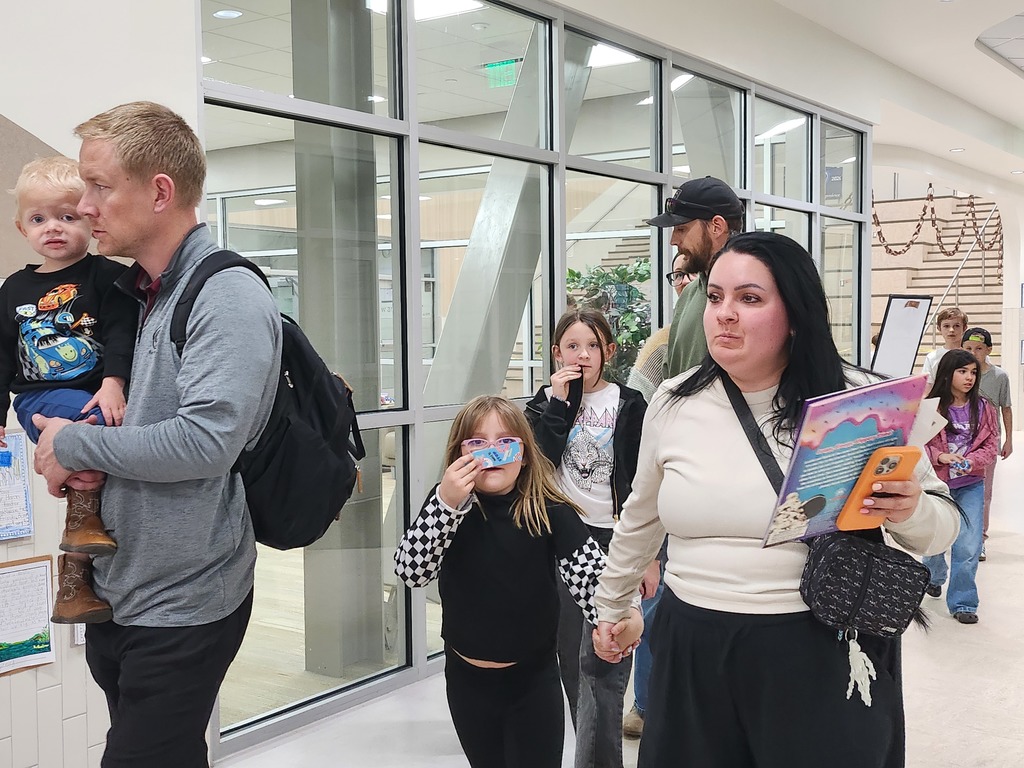 Parents and children walking through a brightly lit school corridor. A woman in the foreground carries a colorful book and a smartphone, while a young girl in a checkered sleeve shirt holds a blue ticket to her face.