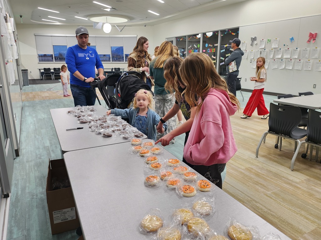 A long table in a school hallway filled with individually wrapped treats, including chocolate cookies and cookies with orange frosting. A young girl in a denim jacket reaches for a treat while parents and other children stand nearby.