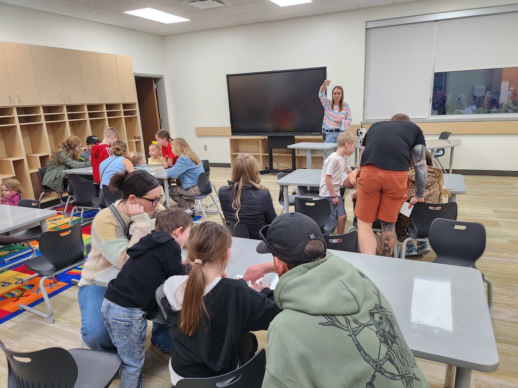 A classroom scene where families and children are seated at tables participating in a hands-on activity. A teacher in a striped sweater stands near a large screen, holding up a small object for the class to see.