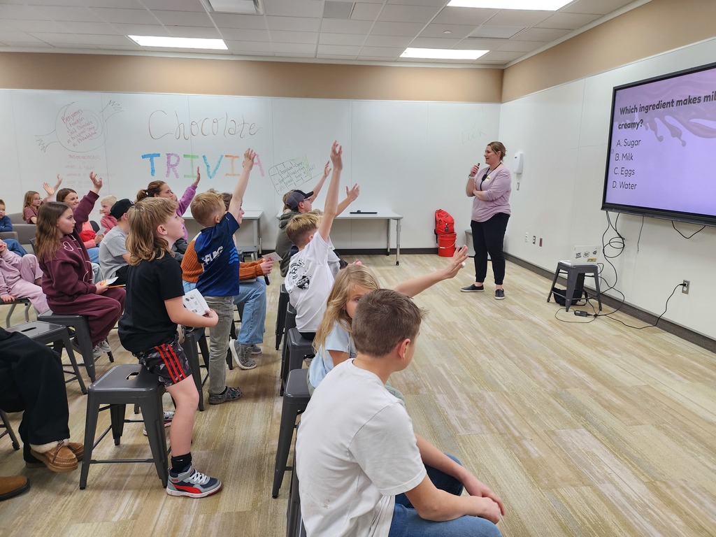 A classroom with a whiteboard that says "Chocolate Trivia" in colorful letters. A teacher stands at the front while students raise their hands to answer a multiple-choice question on a screen: "Which ingredient makes milk chocolate creamy? A. Sugar, B. Milk, C. Eggs, D. Water."