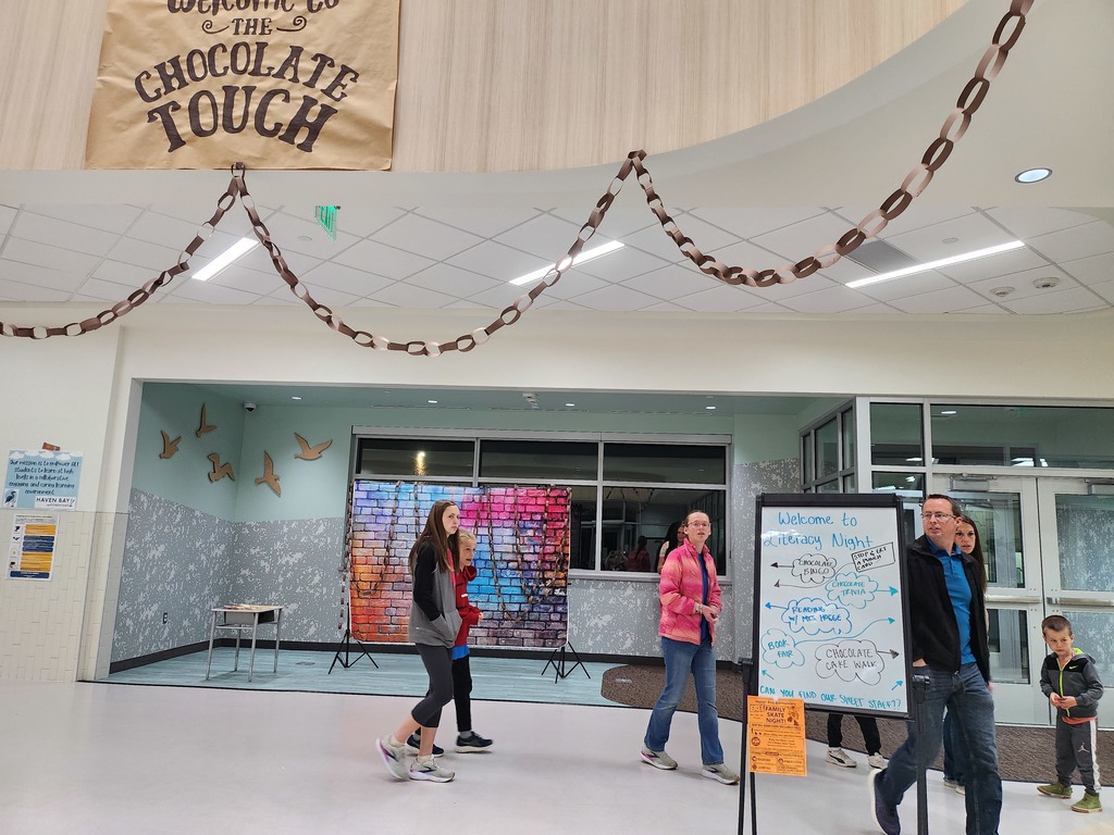 A school lobby decorated with a paper chain and a large brown paper sign that reads "Welcome to the Chocolate Touch." A whiteboard lists activities: "Welcome to Literacy Night," "Chocolate Bingo," "Chocolate Trivia," "Reading with Mrs. Hogge," "Book Fair," and "Chocolate Cake Walk."