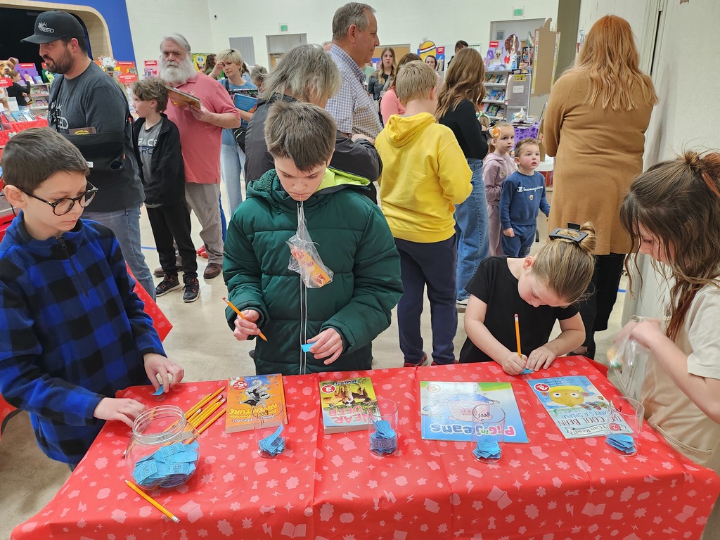 A busy "Literacy Night" event where children are filling out blue raffle tickets at a table covered in a red star-patterned cloth. Books on the table include "A Crabby Adventure," "Bear Grylls Deep Waters," "Pig in Jeans," and "The Cool Bean."