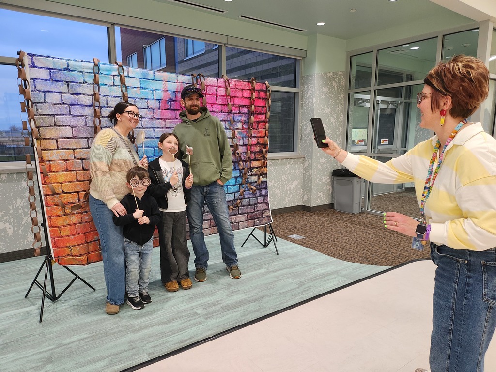 A school staff member uses a smartphone to take a photo of a family of four posing in front of a colorful brick-patterned backdrop decorated with paper chains. The children are holding up photo booth props on sticks.