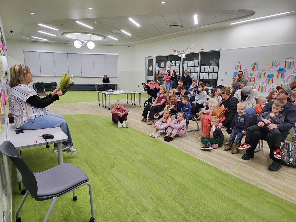 Mrs. Hogge sits on a table in a school common area, reading a book aloud to a group of children and parents seated on a green carpet. A banner in the background reads "Shine Bright," and the wall features a display of colorful paper crafts.