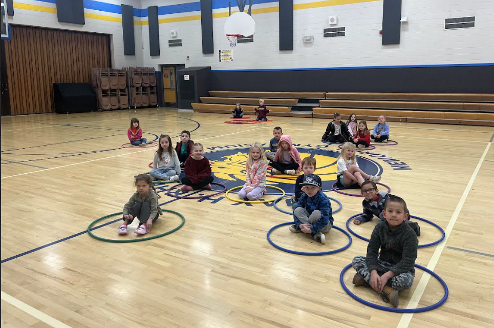 A group of kindergarten students in a gym, each sitting cross-legged inside their own individual hula hoop. They are listening attentively on the basketball court, which features a large lion mascot logo. The hula hoops are various colors like blue, yellow, and green, spaced out across the wooden floor.