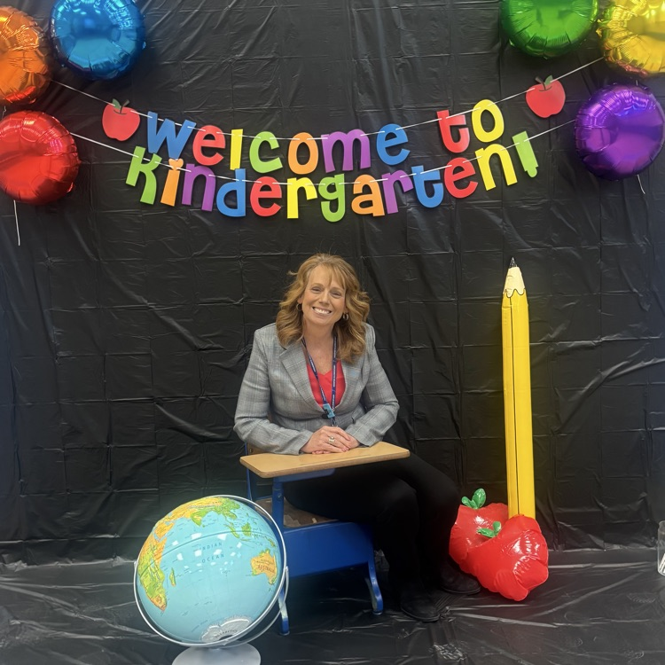Mrs Breeding sitting in front of black background, colored balloons, and  banner that says Welcome to Kindergarten
