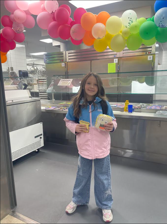 A girl with long brown hair smiles in a cafeteria setting, wearing a pink and blue jacket and wide-leg jeans. She holds two plush toys that look like slices of bread with faces. A large arch of pink, orange, and yellow balloons hangs above the stainless steel kitchen equipment.