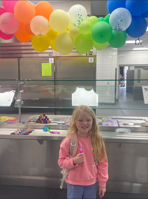 A young girl with long blonde hair and a pink sweatshirt stands in a school cafeteria. She is holding a small pink toy. Above her is a vibrant arch of orange, yellow, and green balloons. Behind her is a stainless steel serving station with various small prizes like Rubik's cubes and stickers. A sign on the wall is partially visible with the letters "NSB".