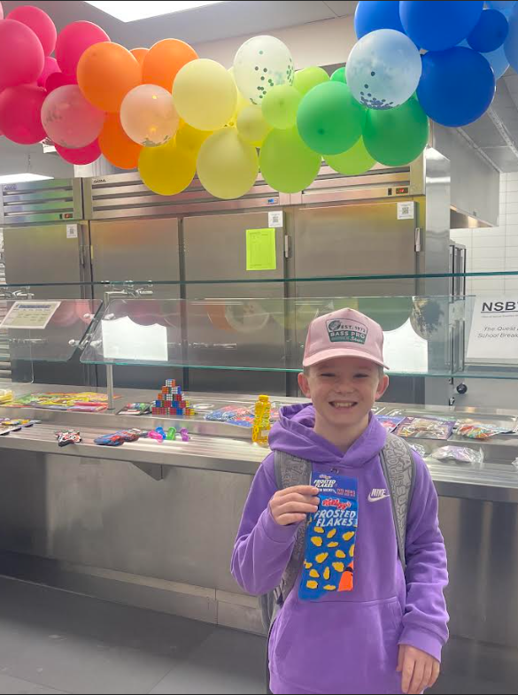 A smiling boy wearing a purple hoodie and a pink "Bass Pro Shops" hat holds up a pair of blue socks featuring the "Kellogg’s Frosted Flakes" logo and mascot, Tony the Tiger. He stands in front of a cafeteria serving line decorated with rainbow balloons. A sign to the right reads "NSBW - The Quest for School Breakfast."