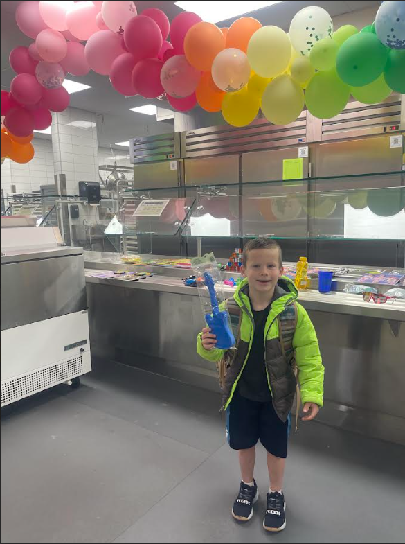 A young boy in a bright neon green and brown vest over a black shirt holds a blue toy guitar still in its plastic packaging. He is standing in a kitchen or cafeteria area under a large rainbow balloon display. The background features industrial stainless steel refrigerators and a counter with various small toys.