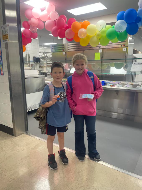Two children, a boy in a blue t-shirt and a girl in a bright pink hoodie, stand together in a cafeteria doorway. The boy holds a small orange toy, and the girl holds a packaged blue item. A large rainbow balloon arch spans the ceiling behind them. To the right, a sign reads "NSBW - The Quest for School Breakfast."