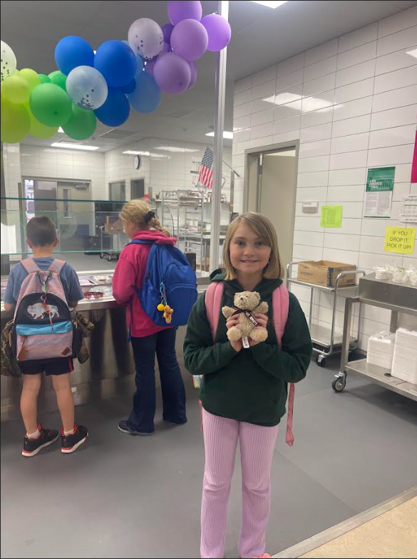 A young girl with blonde hair smiles while holding a small tan teddy bear with a dark ribbon. She is wearing a dark green hoodie and pink ribbed pants. In the background, two other children stand near a metal counter decorated with a rainbow balloon arch. An American flag and a yellow sign that reads "IF YOU DROP IT, PICK IT UP!" are visible on the white-tiled wall.